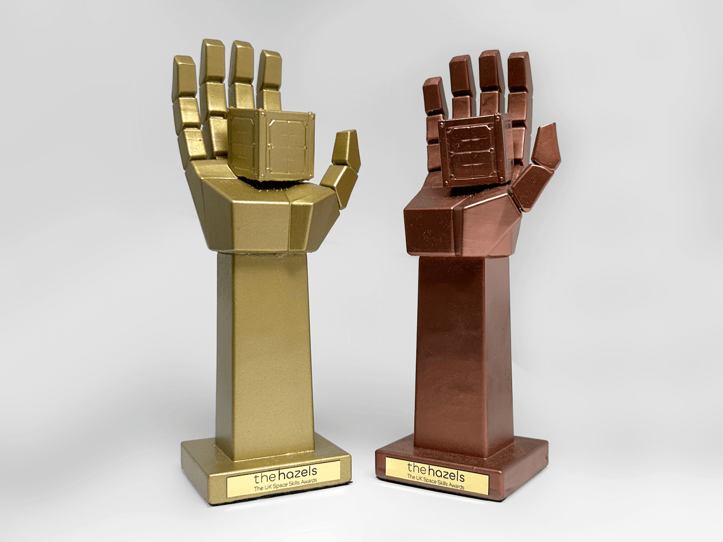 Two custom 3D-printed hand-shaped trophies on stands, one metallic gold and one metallic bronze, each featuring a blocky, geometric design. The stands are labeled with a gold plaque reading 'thehazels'.