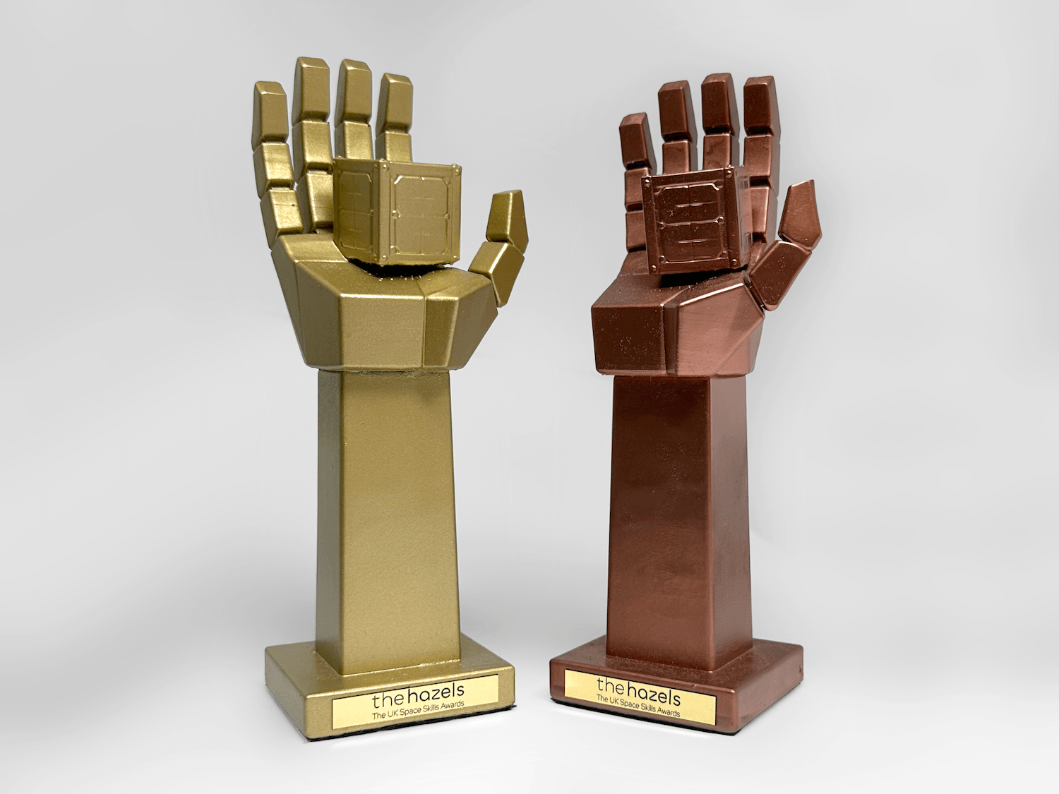 Two custom 3D-printed hand-shaped trophies on stands, one metallic gold and one metallic bronze, each featuring a blocky, geometric design. The stands are labeled with a gold plaque reading 'thehazels'.