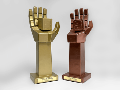 Two custom 3D-printed hand-shaped trophies on stands, one metallic gold and one metallic bronze, each featuring a blocky, geometric design. The stands are labeled with a gold plaque reading 'thehazels'.