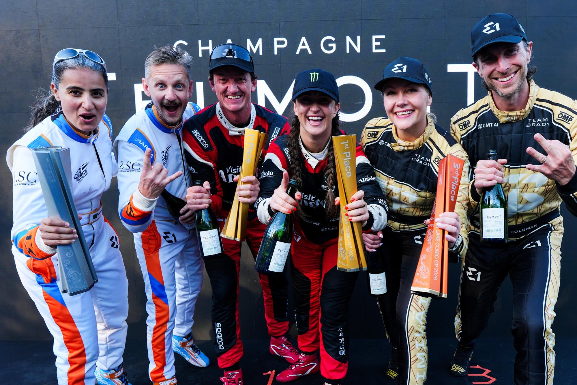 Three winning teams at the E1 Lake Como GP 2026 celebrating in racing overalls, holding Champagne Telmont bottles and silver, gold "1st Place", and orange "3rd Place" trophies.