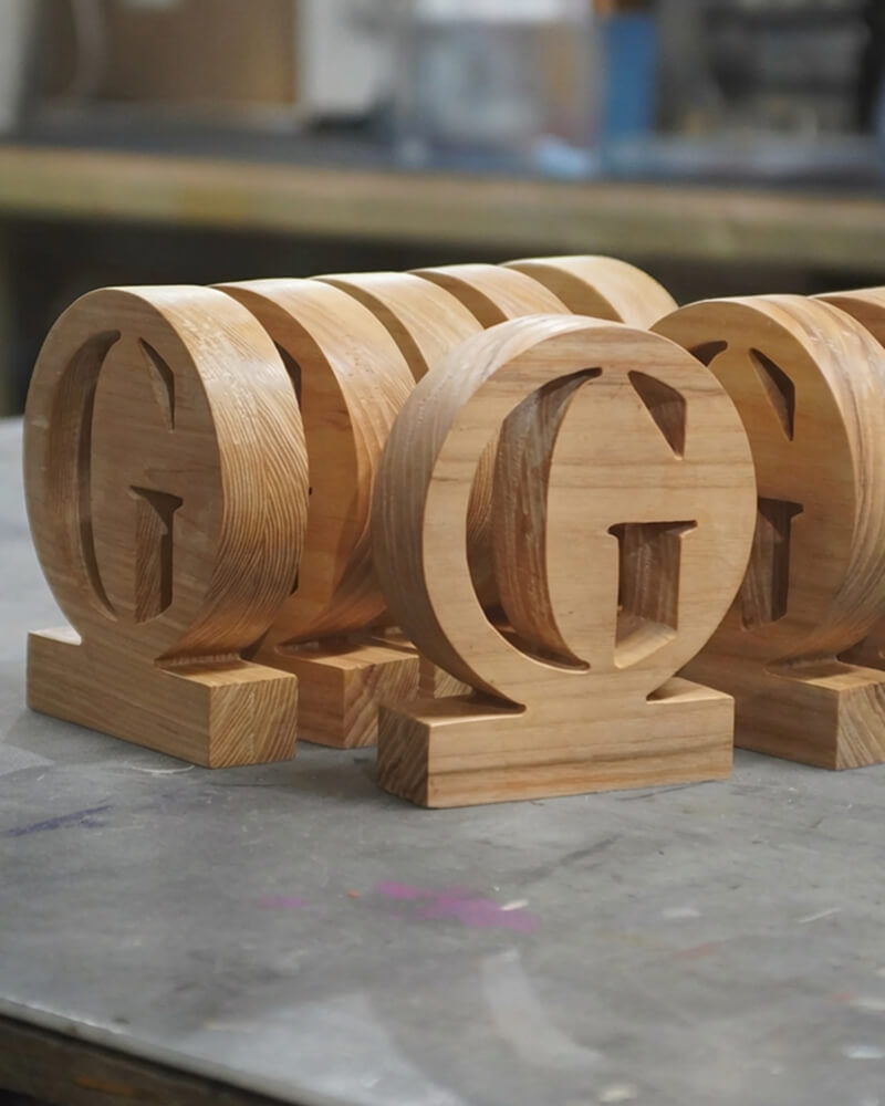 Wooden trophies for the Guardian carved as monogram letters "G" on rectangular bases, several arranged together on a workshop table.