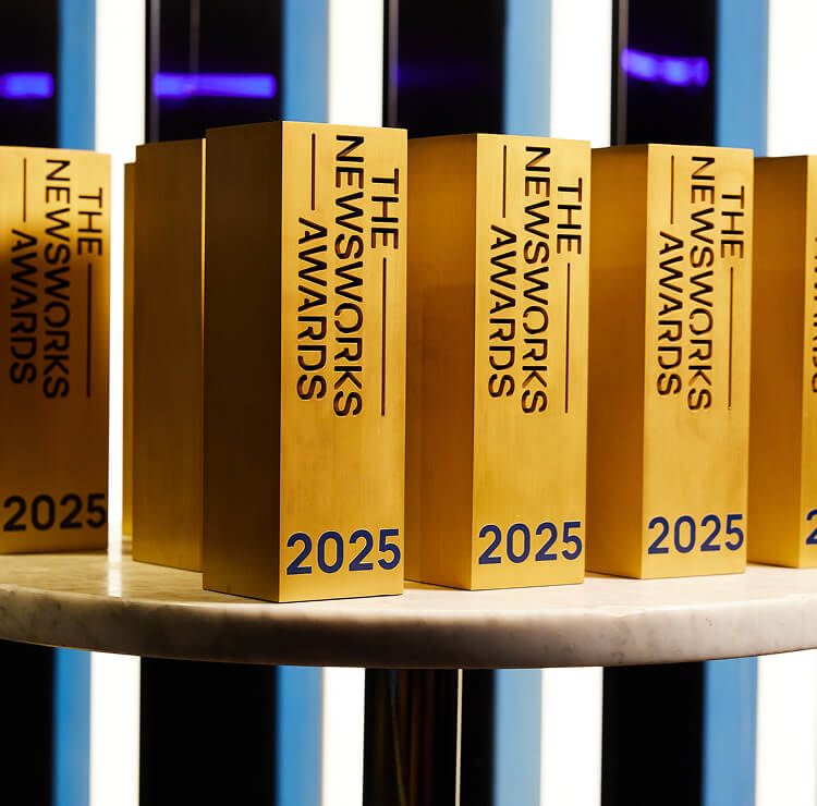 Row of tall gold rectangular trophies engraved "The Newsworks Awards, 2025", on a white marble table against a striped backdrop.