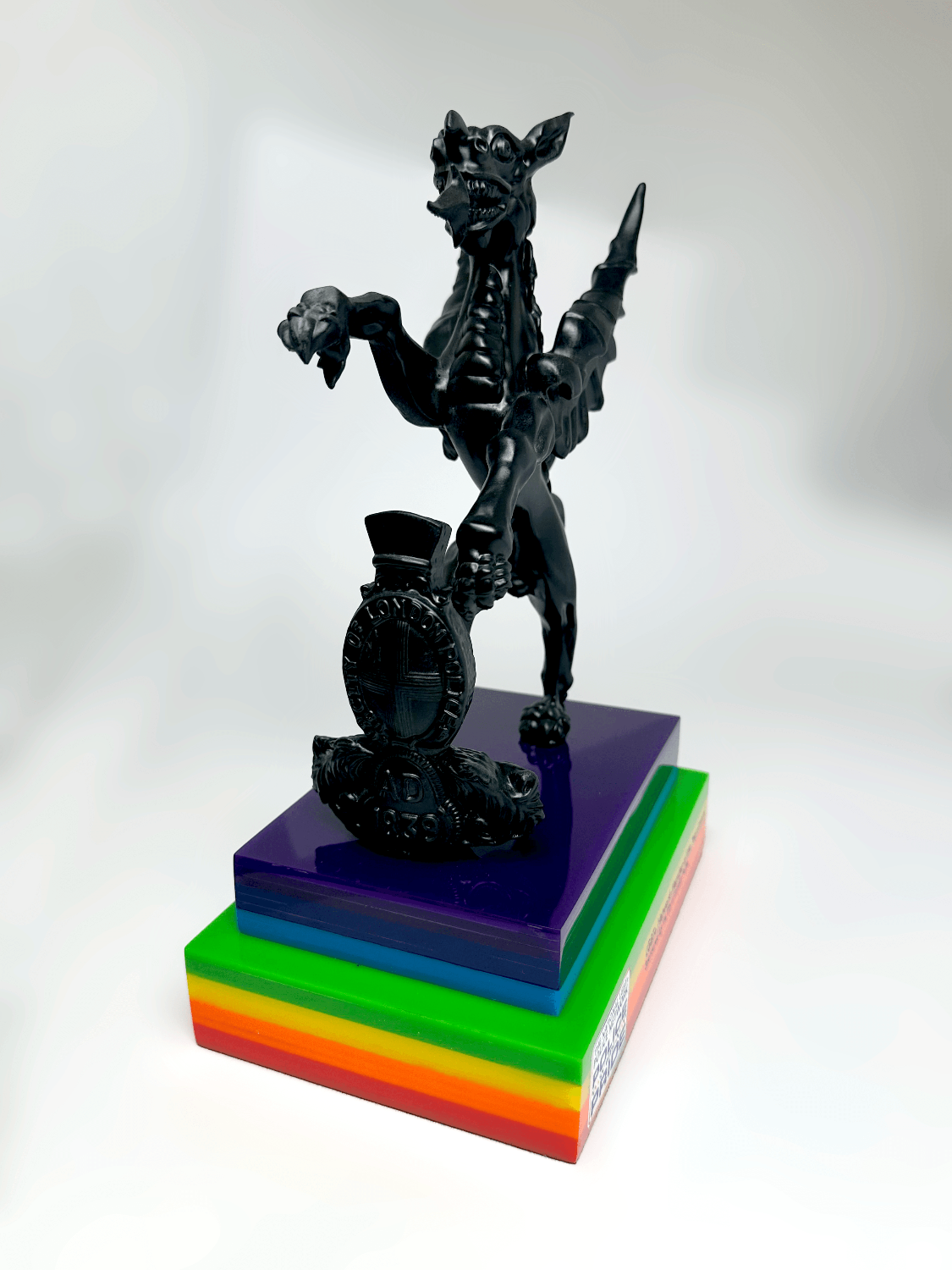 City of London Police and National LGBT+ Police Network for Pride Week griffin figurine standing on a tiered rainbow-colored acrylic base against a white studio background.