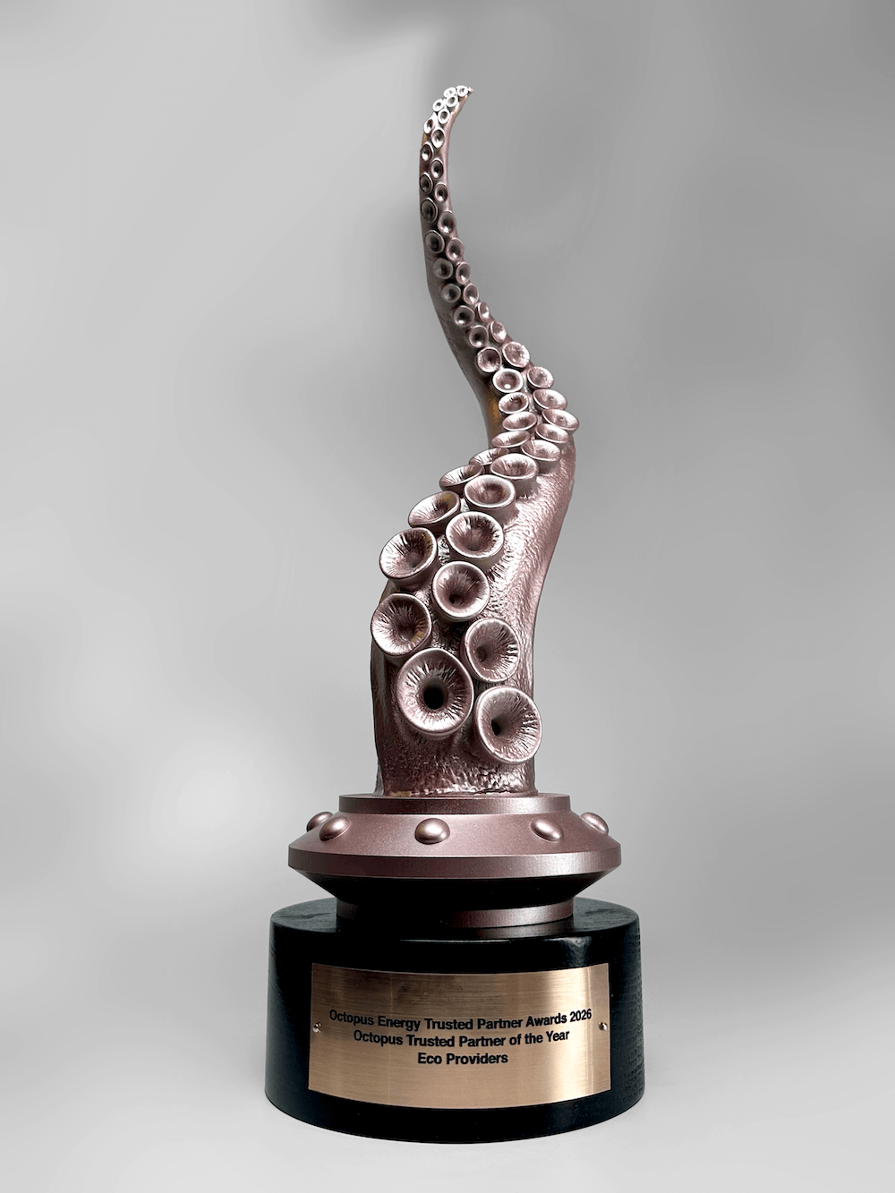 An octopus tentacle sculpture mounted on a round black pedestal with a gold-toned plaque. The sculpture has a metallic rose-gold or coppery finish with detailed suction cups, set against a neutral gray studio background.