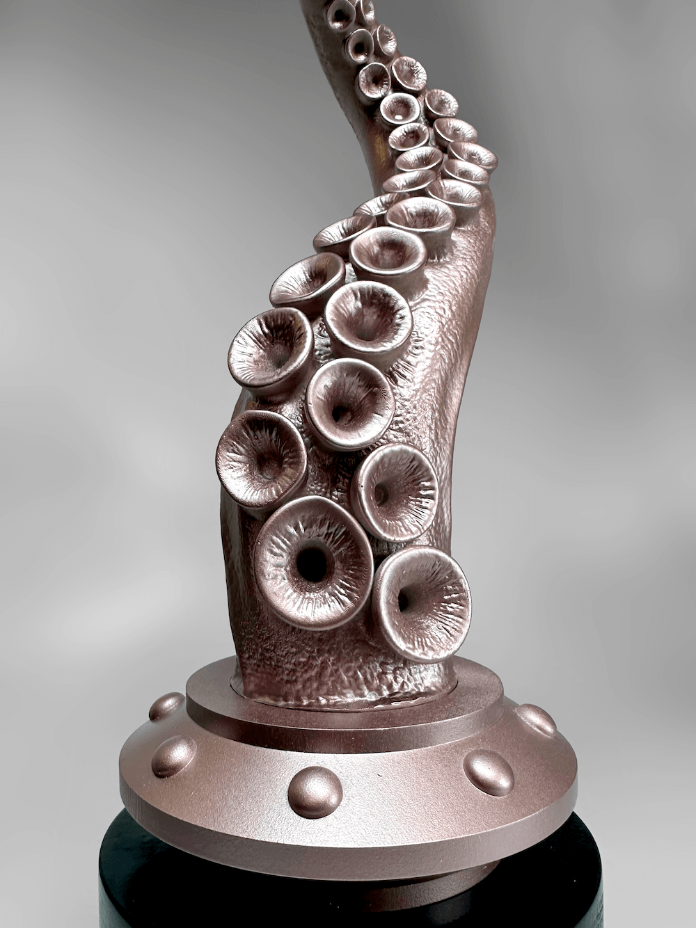 A close up metallic bronze octopus tentacle sculpture with suction cups, mounted on a round base, photographed against a soft gray gradient background.