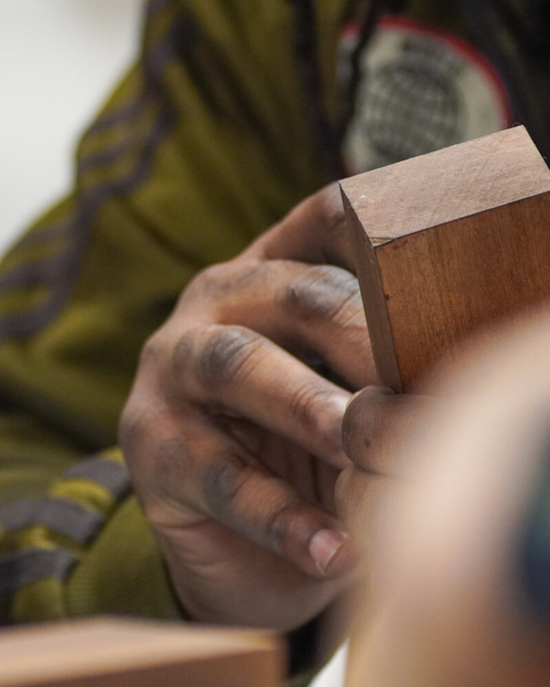 Close-up of a craftsperson's hands sanding a small wooden award base, wearing an olive-green sleeve.